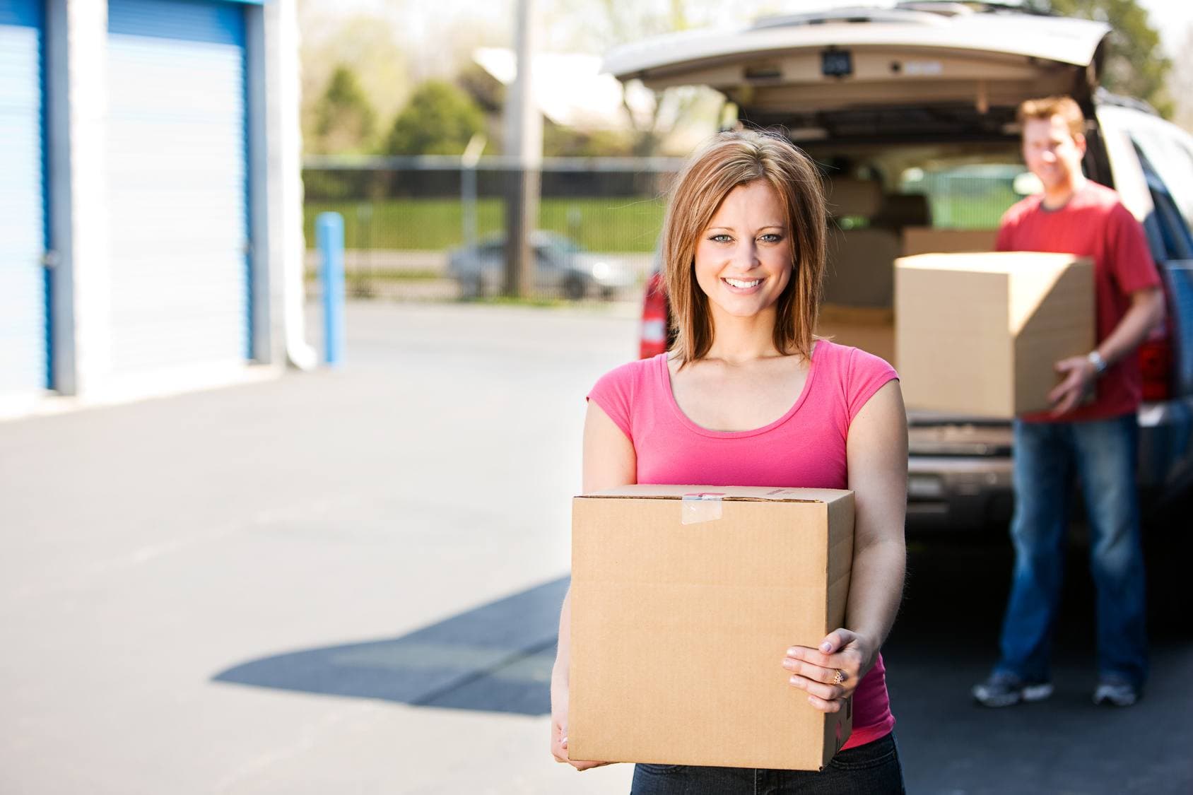 Smiling woman holding a cardboard box while a man unloads an SUV at storage units.