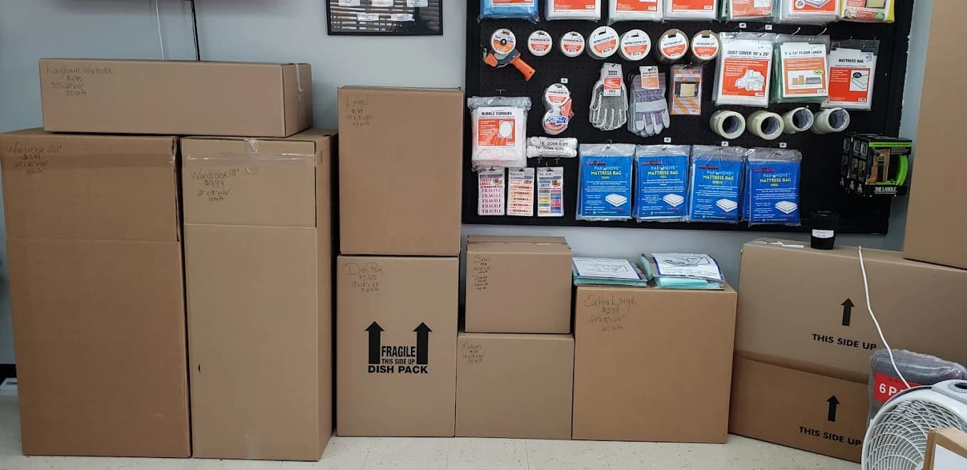 Cardboard moving boxes stacked below a pegboard wall displaying various packing and shipping supplies.
